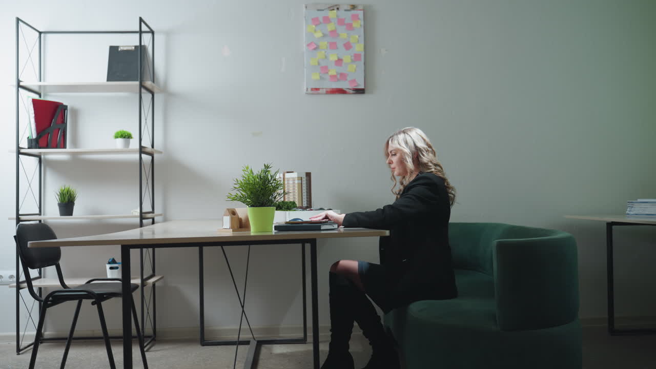 Real estate saleswoman sits down at desk, pulls laptop forward, opens it to begin workday in bright modern office with potted plant, model building, organized shelves, and green couch in background