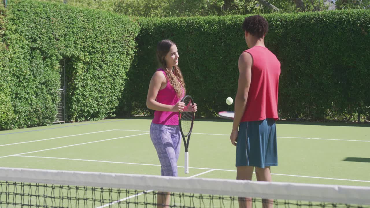 Coppia felice di etnia mista con racchette da tennis e palla che conversano in giardino in una giornata di sole