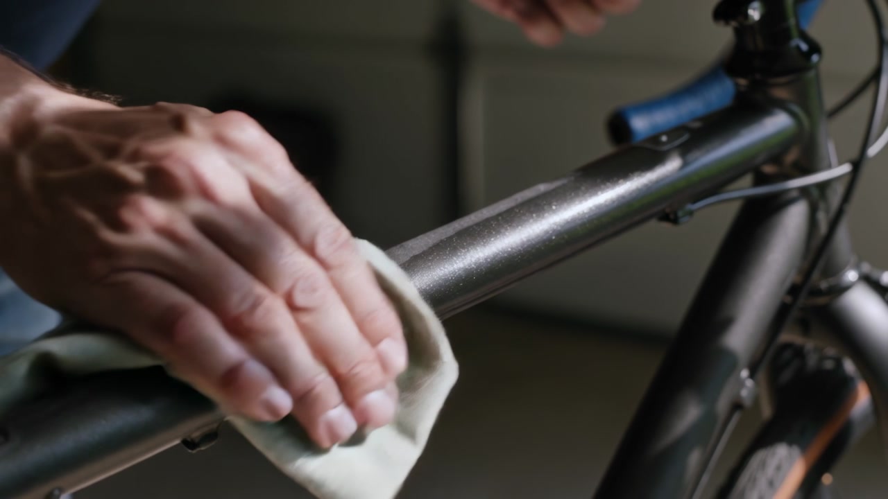Noticing dirt, person's hands wiping carefully bicycle top tube in garage with pale cloth