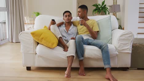 smiling mixed race gay male couple sitting on sofa embracing watching tv