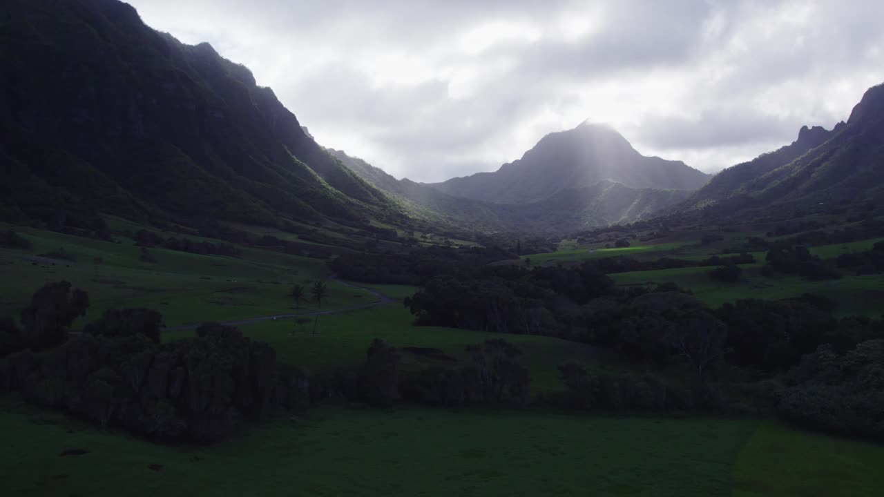 Aerial view of a dramatic Hawaiian valley as sunlight streams through heavy clouds, casting a soft glow over green fields and rugged mountain slopes.