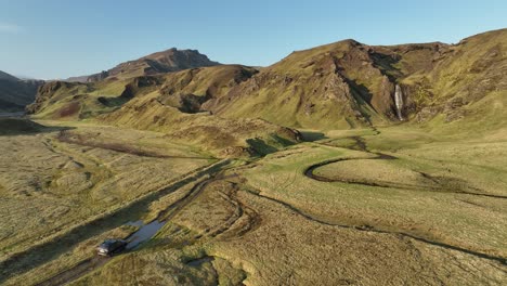 aerial view of an off-road vehicle driving through a remote valley in northern iceland, with rugged hills, winding streams, and a distant waterfall