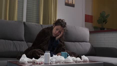 young woman sick on the sofa at home, blowing her nose with handkerchiefs. pills, tissues and a cup on the table