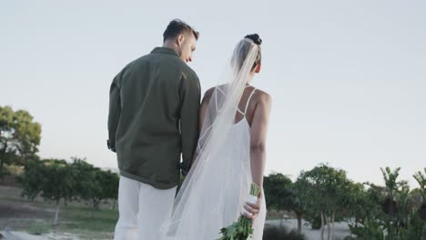 Rear-view-of-happy-diverse-bride-and-groom-walking-holding-hands-at-beach-wedding,-in-slow-motion