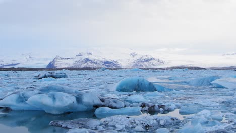 Das-Schwarze,-Bläulich-Weiße-Eis-Auf-Einem-See-In-Island-An-Den-Schneebedeckten-Bergen---Weite-Pfanne