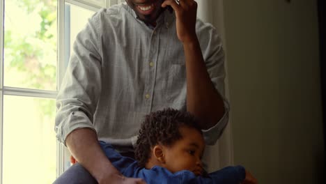 Front-view-of-young-black-father-talking-on-mobile-phone-with-baby-in-a-comfortable-home-4k