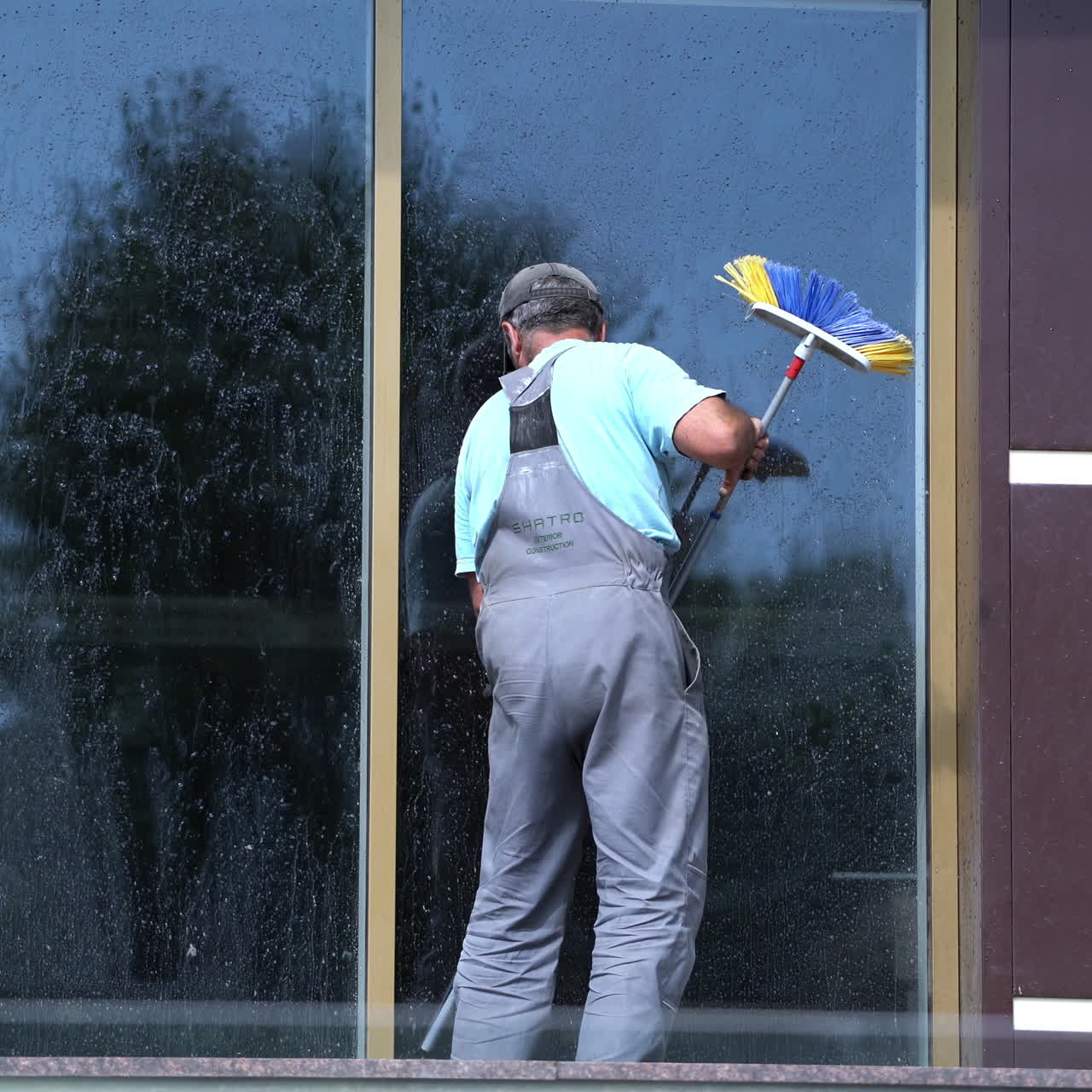 Man washes office window. Back view of a male worker who is cleaning window with a mop outdoors. Washing glass window of office building. Building cleaning services.