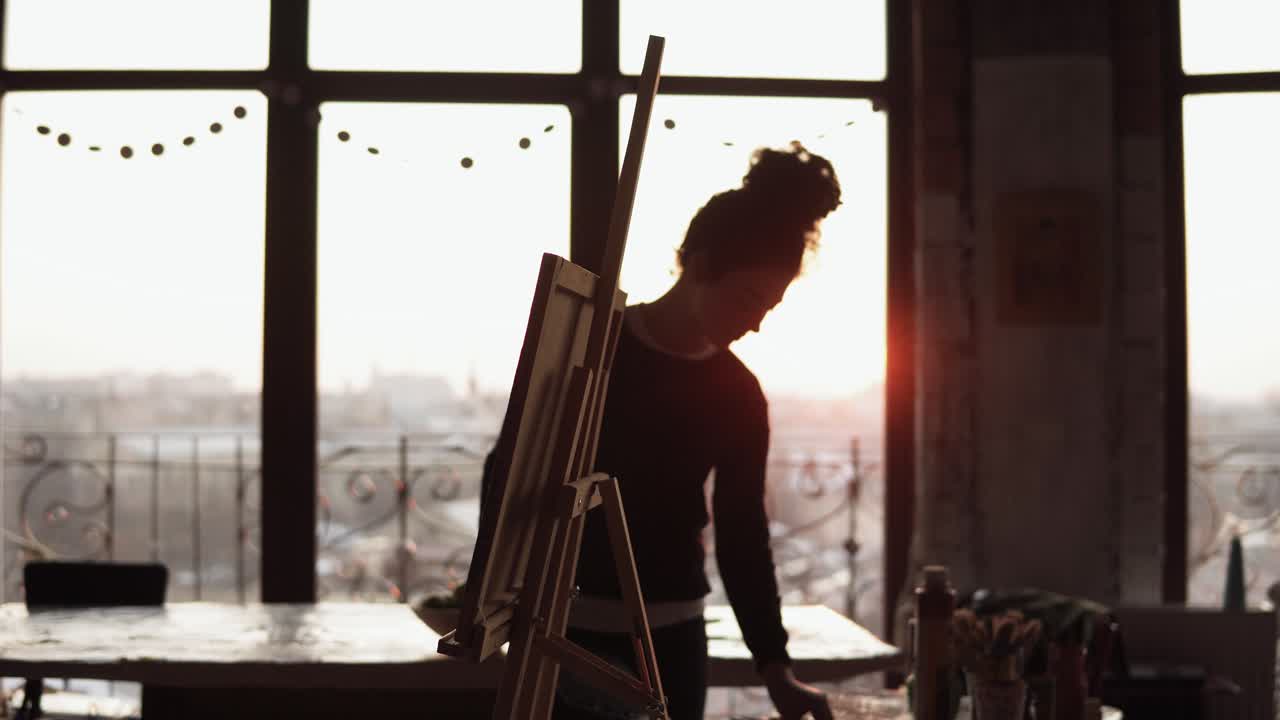 una joven artista profesional inspirada pone cancas en un caballete, toma una paleta con pintura y comienza a dibujar.