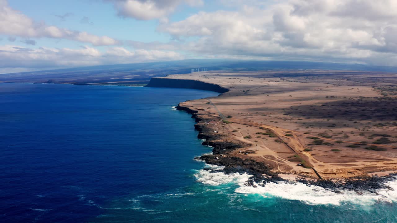 A sweeping coastal landscape where deep blue waters slam into vertical volcanic cliffs, while dry inland tracks carve through sparse brush toward a vast elevated plateau under textured skies