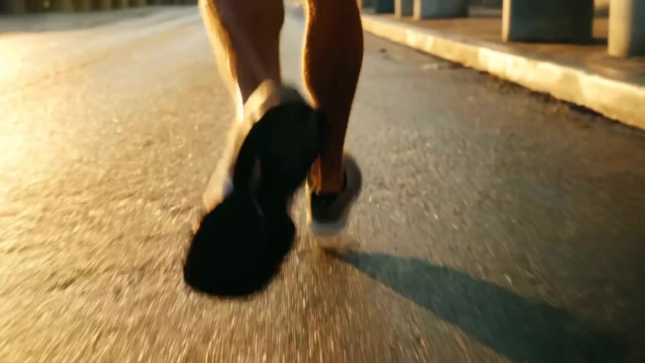 Close-up sudut rendah kaki bagian bawah dan sepatu kets seorang pelari yang berlari di sepanjang jalan kota yang diterangi matahari saat jam emas. Cahaya samping yang hangat membentuk bayangan panjang di aspal