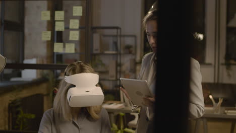 woman emplooyee at desk in the office wearing virtual reality glasses while her coworker is controlling her with tablet 3