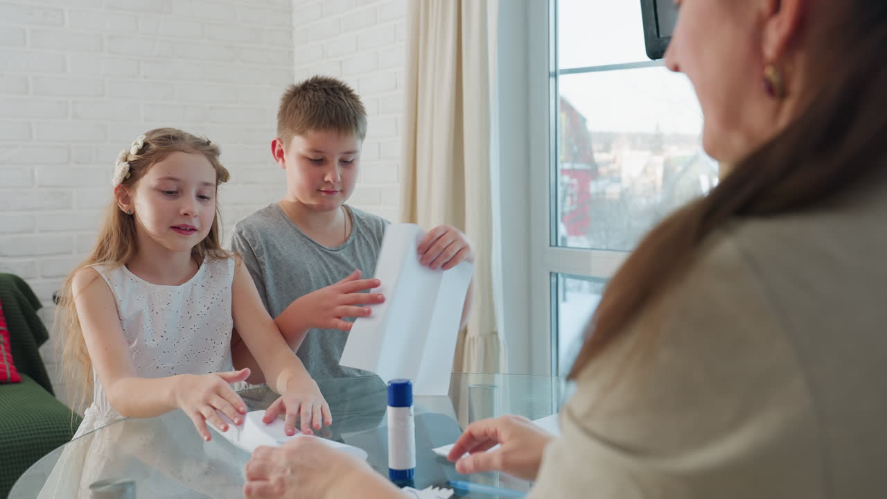 kids sitting at table in front of their mom, holding paper and folding it trying to create something, family crafting activity, kids and mom engaged in creative fun, happy moment together