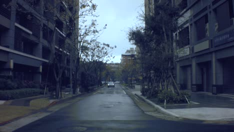 Noch-Immer-Blick-Auf-Ein-Schickes-Geschäftsviertel-Mit-Blick-Auf-Eine-Nasse,-Feuchte-Betonstraße-Mit-Einem-Fahrzeug,-Das-An-Einem-Trüben,-Hellen,-Regnerischen-Tag-Mit-Kleinen-Bäumen-In-Die-Tiefgarage-Einbiegt