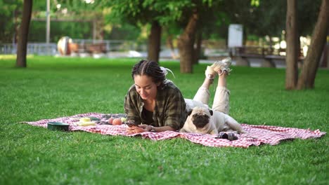 mujer acostada al aire libre en el césped de césped usando su teléfono inteligente y lindo pequeño pug está acostado cerca