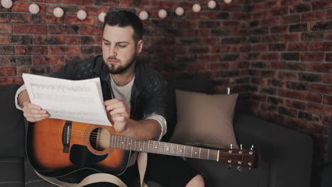 young musician man looking at sheet music and playing guitar at home