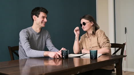 man and blind woman in sunglasses sitting at table at home talking 1