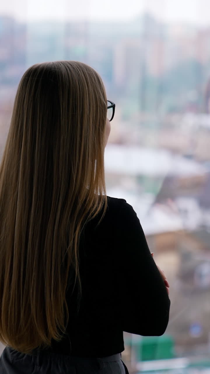 Attractive business lady in glasses standing at window. Long-haired woman looks through the window on a city landscape. Rear view. Vertical video