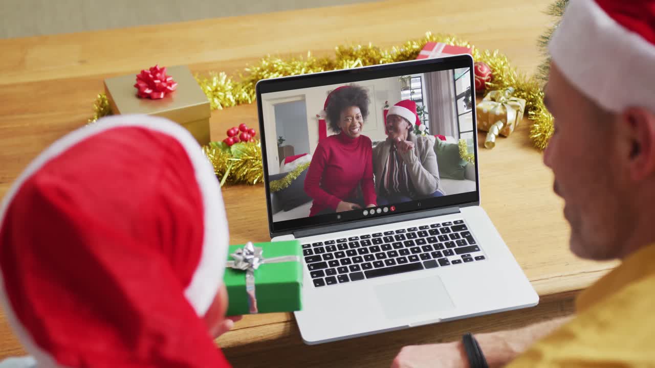 padre y hijo caucásicos con sombreros de santa usando una computadora portátil para una videollamada de navidad con la familia en la pantalla