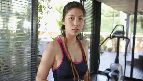 serious asian woman with skipping rope on neck looking at camera in sunny home gym, slow motion