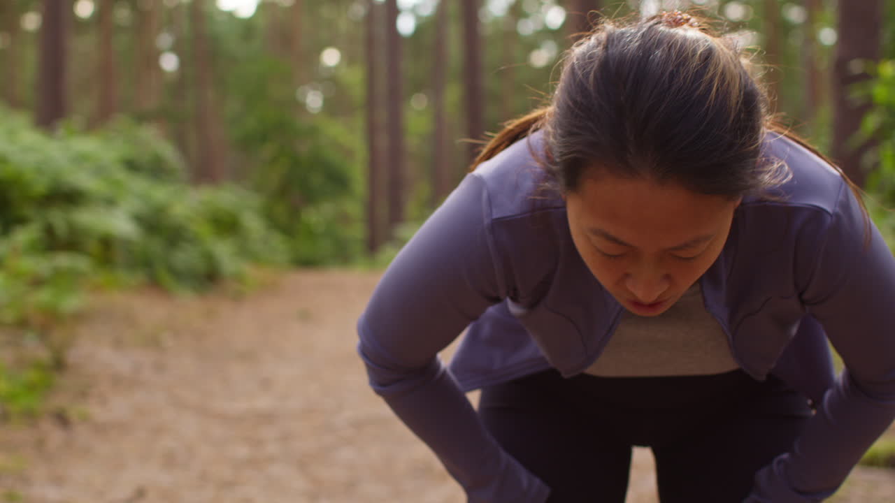 피곤한 중성인 여성이 야외에서 운동을 하고 숲을 가로질러 달리고 숨을 쉬기 위해 멈추고 실시간으로 습니다.