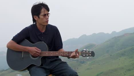 asian romantic guy with glasses and long hair playing a guitar ballad in mountains