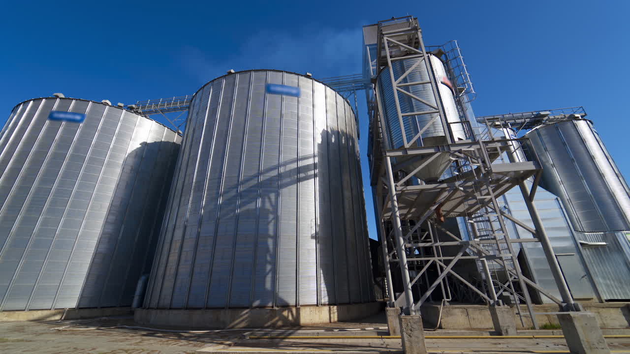 Modern factory for storage crop. Metal grain elevators on agricultural complex. Exterior of industrial plant under blue sky.