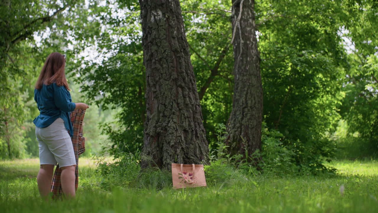 Lady picks up picnic blanket from grass preparing to fold after resting under tall trees in green park, wearing blue shirt and white shorts, enjoying peaceful outdoor summer day surrounded by sunlight