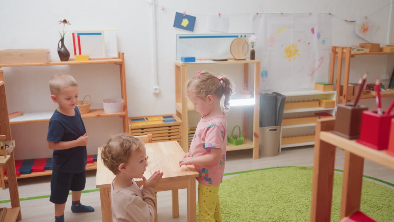 Unos niños en una clase Montessori observan un objeto sobre una mesa mientras un chico con estrabismo se inclina hacia atrás jugando, mientras sus amigos observan con entusiasmo y curiosidad durante una actividad grupal cerca de unas estanterías.