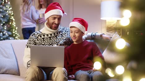 Portrait-Of-Loving-Dad-In-Sana-Hat-With-Teen-Son-Sitting-At-Home-Browsing-On-Laptop-Spending-Holiday-Time-Watching-Xmas-Movie