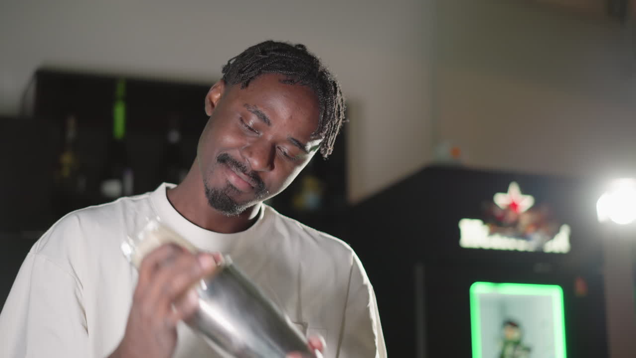 Young man shaking cocktail shaker, focused expression in modern bar with Heineken logo blurred in background. Perfect for videos featuring bartenders, cocktail preparation, and mixology moments