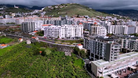 sweeping shot of coastal hotels in madeira , revealing the sea at the end of the shot , with the lush green mountains in the background with loaming low clouds