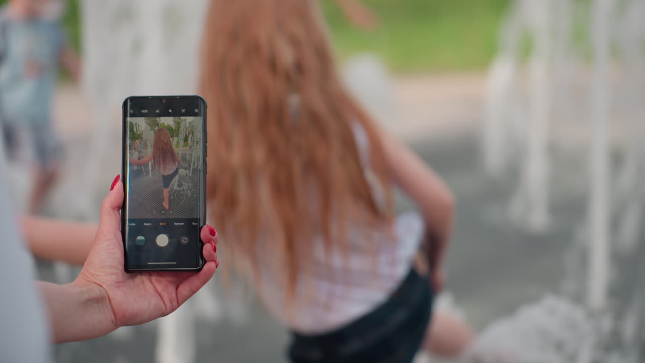 toddler playing in fountain captured on phone camera, parent hand holding device, water jets surrounding child, summer park background, joyful movement and candid family outdoor fun