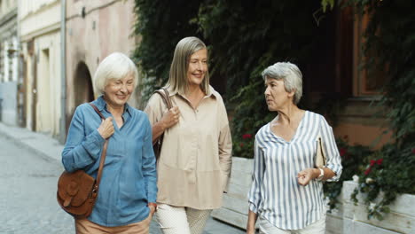 Three-Cheerful-Senior-Women-On-Retirement-Walking-The-Old-City-Street-And-Laughing-While-Talking