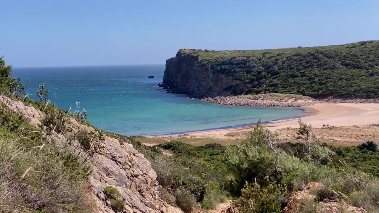 Vista paesaggistica costiera di una baia panoramica con acqua turchese, una spiaggia sabbiosa e scogliere verdi in una giornata di sole.