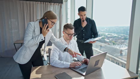 man in white shirt and glasses sits in front of laptop. male and female colleagues standing on both sides of him. woman speaking on the phone.