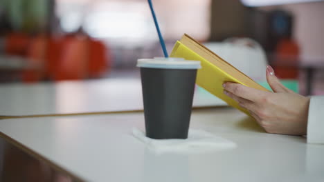 close-up of hands holding a yellow book with polished nails, coffee cup on the table, and blurred background featuring warm lighting and soft tones