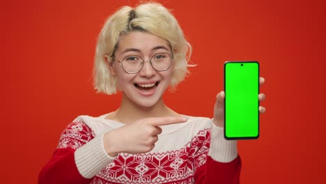 christmas. young asian woman presenting smartphone with green screen forefinger showing swipe gesture in christmas clothes to copy space to advertise products on red background. discounts sale concept