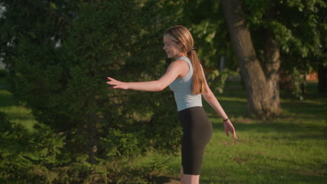 side view of young lady smiling while skating outdoors on sunny day, arms wide for balance, surrounded by lush greenery and trees, near grassy field, with warm sunlight reflecting off her