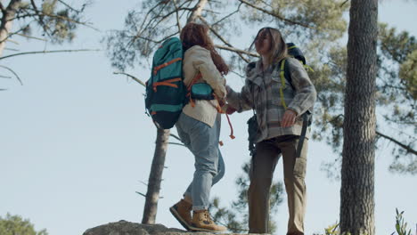 young woman backpacker helping her friend to climb on the top of a stone, then hugging and enjoying the view