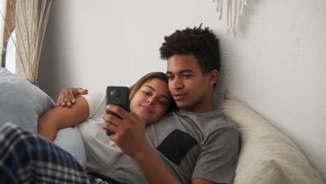 happy multi ethnic young couple relaxing in bedroom with the smartphone embracing each other. slow motion shot