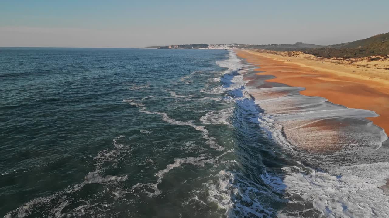 Flygande dolly-framåtfotografering av Atlantens vågor som slår mot den sandiga kusten. Denna natursköna vy visar Praia do Salgado-stranden nära Nazaré, Portugal, en ljus och solig dag.