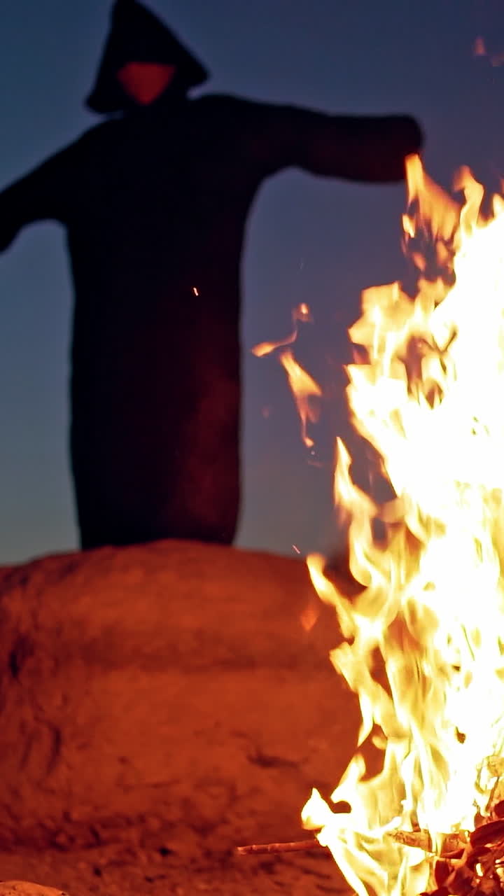 Scary figure in black mantle. Close up of witch stay around the fire in forest. Vertical video