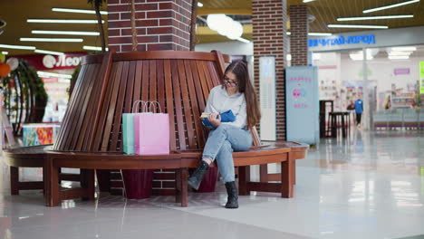 young lady seated on a modern wooden bench in a well-lit mall, leg crossed, engrossed in a book with light reflecting on the shiny mall tiles and shopping bags beside her