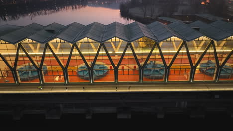 People-walking-and-on-a-bicycle-over-a-bridge-next-to-the-illuminated-tubrine-house-of-the-hydro-power-plant-in-Birsfelden-while-the-colorful-sky-is-reflected-by-the-river-Rhine