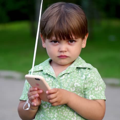 the boy uses a mobile phone holding it in his hands. cute toddler with long eyelashes is busy with a cell phone. the boy is in nature