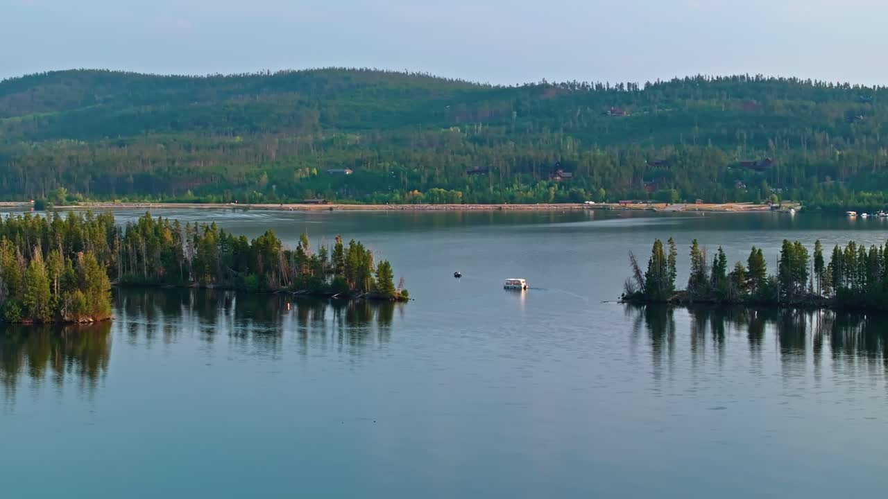 Drone aerial orbit of boat on reflective Grand Lake Colorado surrounded by pine forest and distant mountain range backdrop
