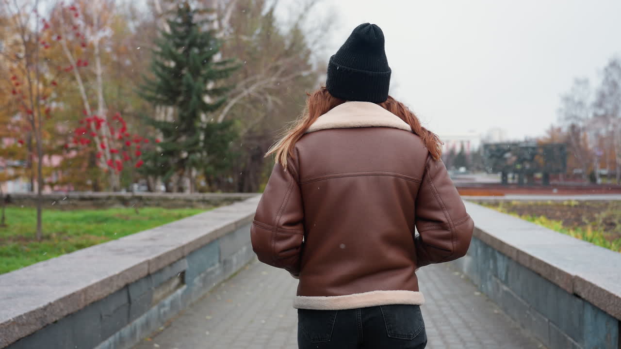 Back shot of student in black cap brown jacket walking slowly with hands in pockets on wide stone pathway through urban park surrounded by pine trees modern buildings and cloudy sky