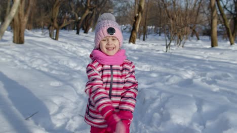 Child-girl-running-on-snowy-road,-fooling-around,-smiling,-looking-at-camera-in-winter-park-forest