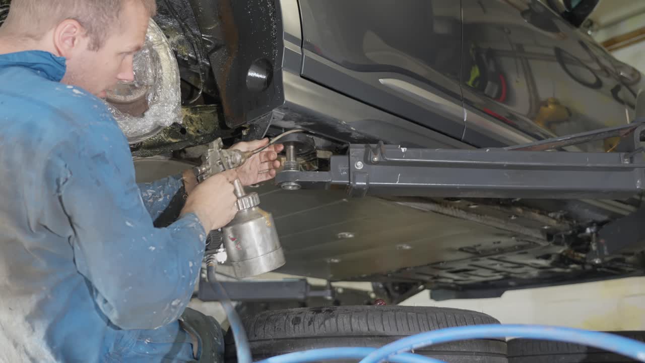 Man in coveralls applying lanolin rustproofing inside car sill and rocker panel cavities with long tube attached to pressurized spray gun