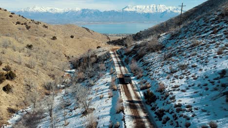 A-truck-driving-on-a-dirt-road-through-a-narrow-canyon-towards-a-valley-with-a-lake-and-snow-capped-mountains